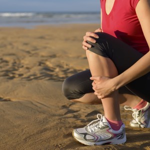 Female runner clutching her shin because of a running injury and inflammation. Tibial periostitis hurt while jogging on beach.